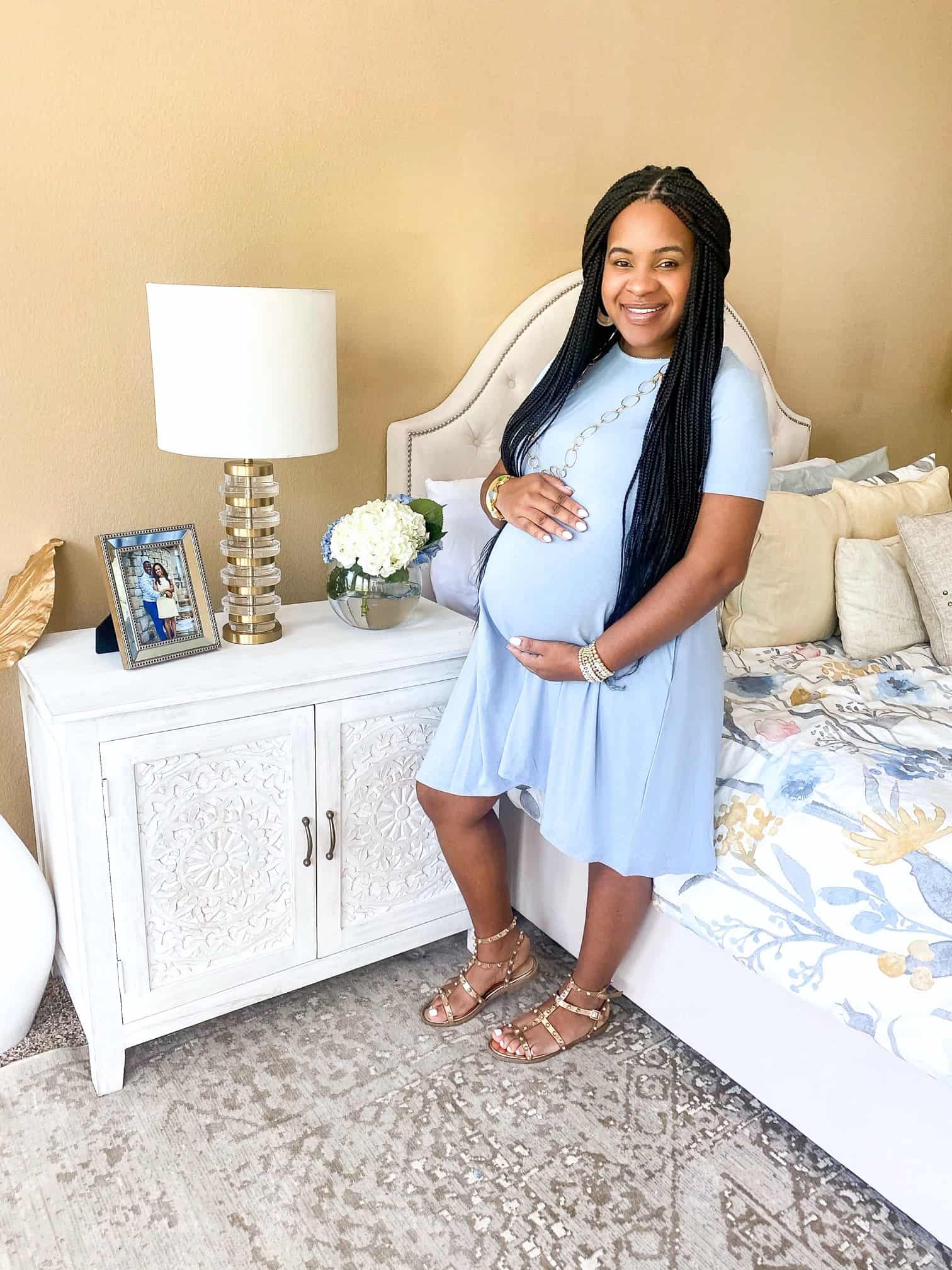 Home Building Experience by popular Dallas lifestyle blog, Glamorous Versatility: image of a woman wearing a blue maternity dress and holding her belly with her hands as she stands next to her bed in her bedroom. 