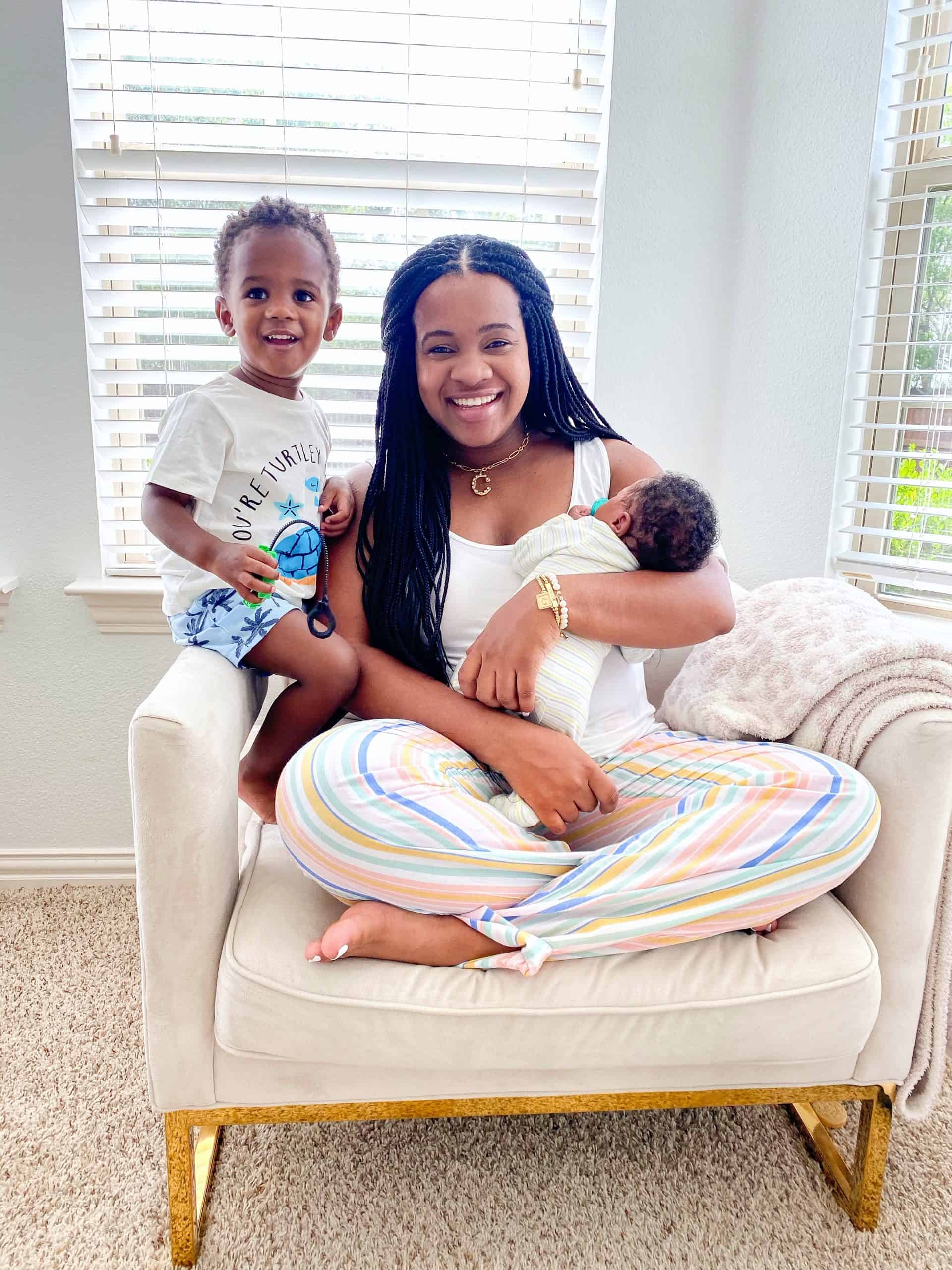 Postpartum Recovery Tips by popular Dallas motherhood blog, Glamorous Versatility: image of a woman wearing multi-color stripe pajama pants and a white tank top and sitting in a cream armchair with her newborn baby boy and toddler aged son. 