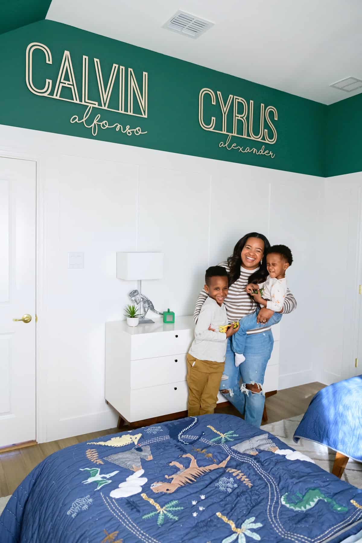 Bedroom Paint Colors by popular Dallas life and style blog, Glamorous Versatility: image of a mom and her two boys standing together in a room decorated with a white dresser, dinosaur bedding, and wall name decals. 