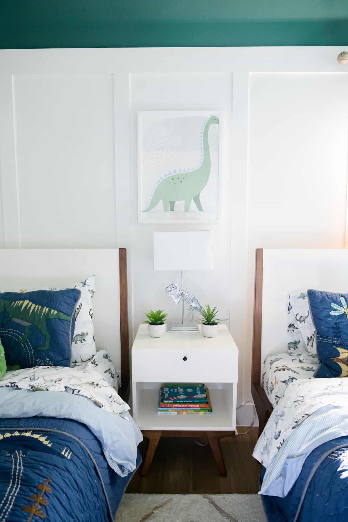 Dinosaur Themed Room by popular Dallas life and style blog, Glamorous Versatility: image of a boys room with a green painted ceiling, dinosaur bedding, framed dinosaur artwork, dinosaur lamp, board and batten walls, and a white night stand. 